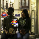 A young man and woman sitting by a window in Madrid, holding an olive plant and a Spanish language book, representing a new life and asylum in Spain, HIGH NEWS.