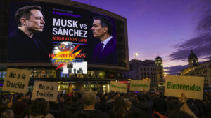 Elon Musk and Pedro Sánchez on a digital billboard in Madrid debating Spain migration law with protesters holding signs.