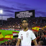 A soccer player on the pitch with a large stadium screen displaying a warning against Islamophobic and xenophobic chants during a match.