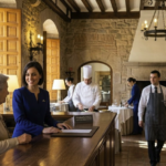 A smiling female receptionist assisting a guest at a Paradores hotel in Spain, with kitchen staff and a waiter in the background, representing permanent job opportunities in Spain.