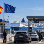 Spanish and EU flags at a port border checkpoint with cars, police officers and a passenger ferry.